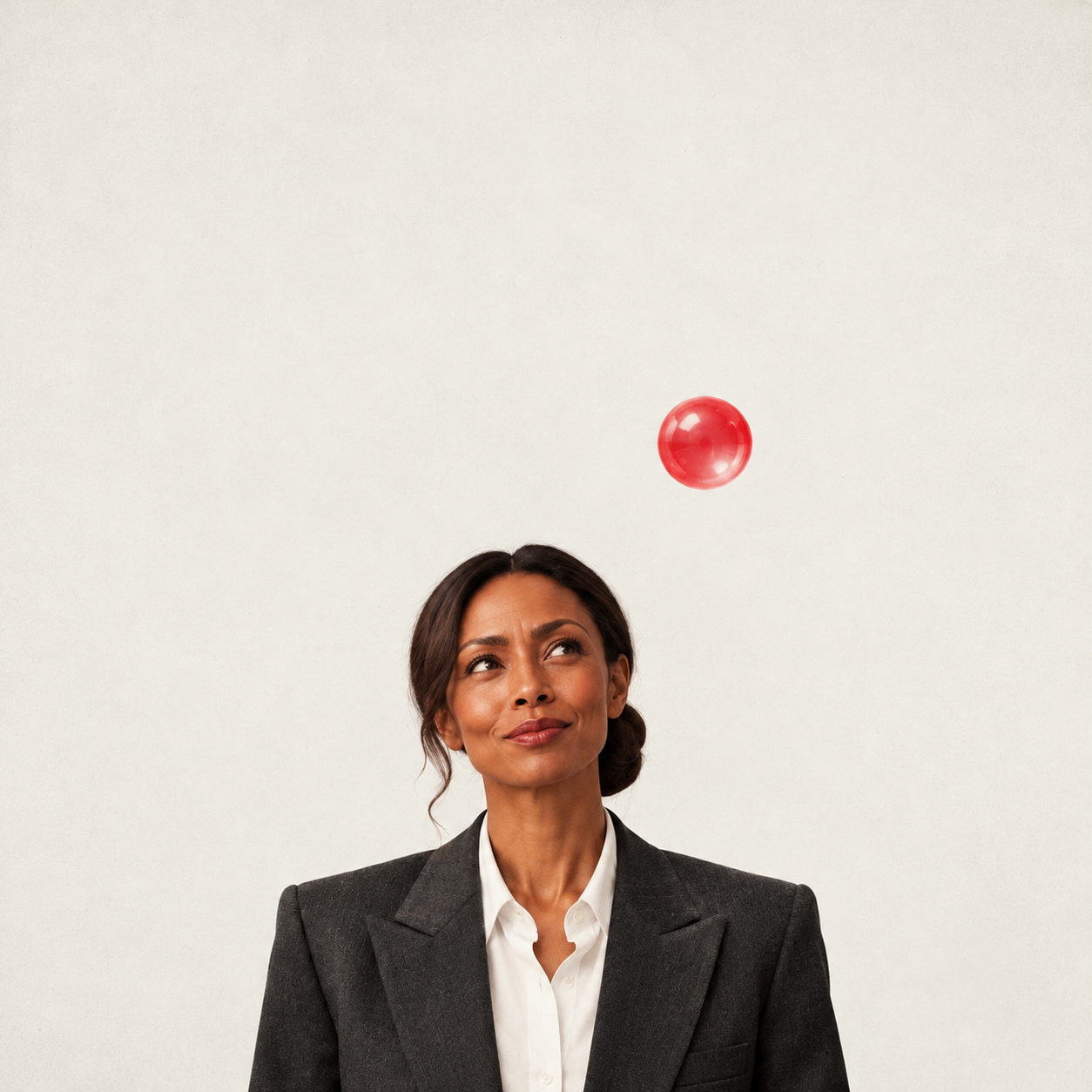 A woman in a charcoal suit looks up at a single red bubble floating against a cream background.